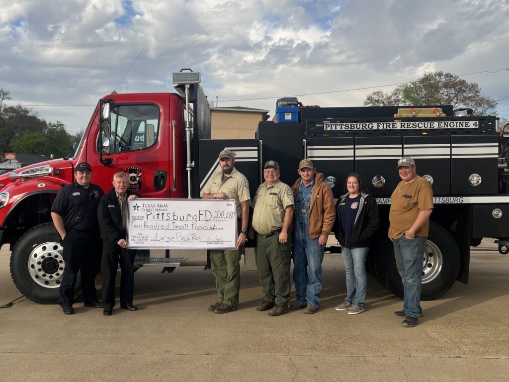 Group photo of Pittsburg Fire Department members standing in front of a red and black fire engine, holding a large check.
