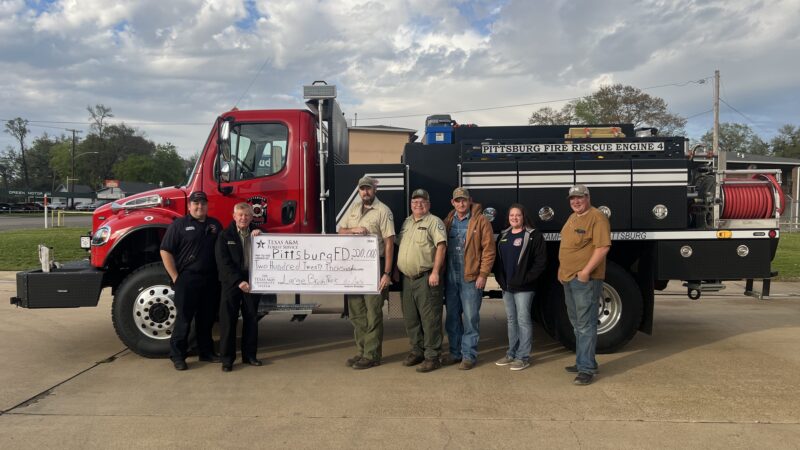 Group photo of Pittsburg Fire Department members standing in front of a red and black fire engine, holding a large check.