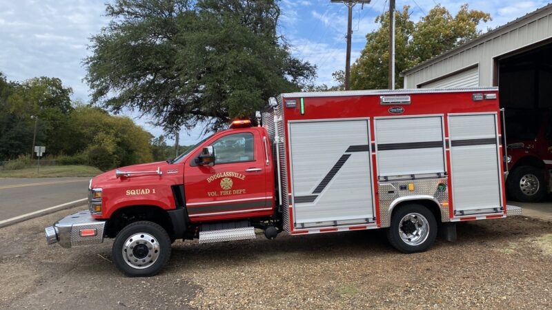 A bright red fire rescue truck labeled "Douglasville Vol. Fire Dept, Squad 1," parked outside a fire station, with a tree and clear sky in the background.