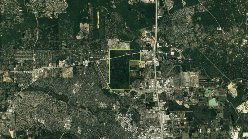 Satellite image of a sprawling urban area with a mix of residential neighborhoods, green spaces, and roads. A major highway runs vertically through the scene, surrounded by dense clusters of houses and some large open green plots.