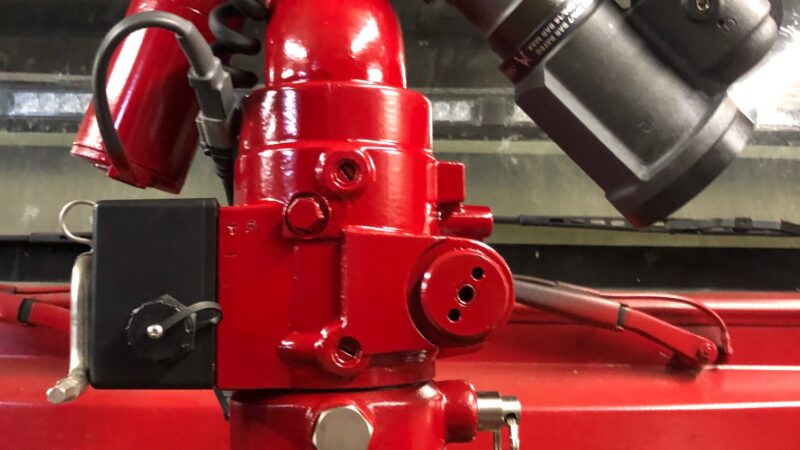 Close-up of a red fire truck nozzle with various attachments and a hose connection, mounted on the vehicle. The nozzle features adjustable joints and control levers, designed for directing water during firefighting operations.