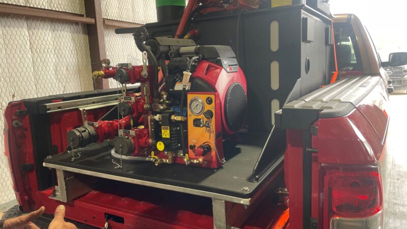A red utility truck with a custom setup in the bed, featuring various tools and machinery, including gauges and hoses.