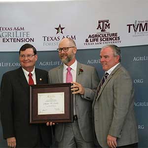 Three men smile in front of backdrop with framed award