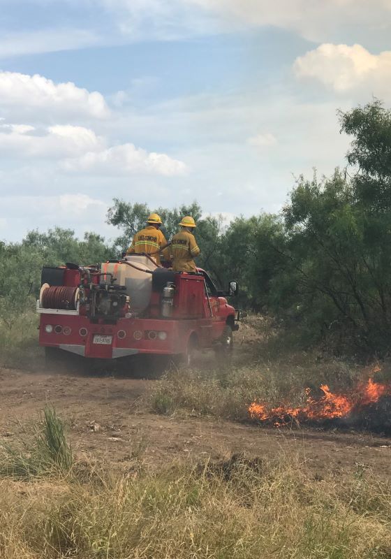 Fire tuck with firefighters standing on top driving through field with flames