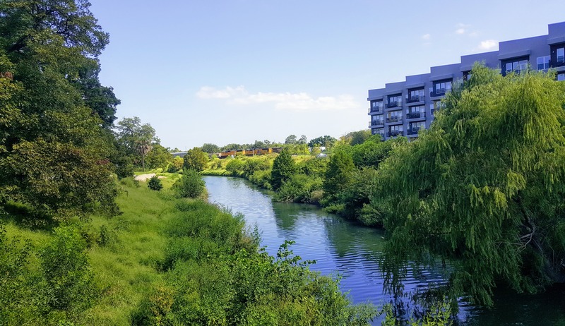 Image of a flowing body of water winding through an urban area, bordered by green vegetation on both banks, with buildings visible in the background.
