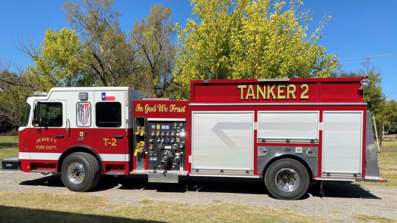 A red Jewett Fire Dept. tanker truck labeled "TANKER 2" with "In God We Trust" displayed, parked on grass with trees and a blue sky.