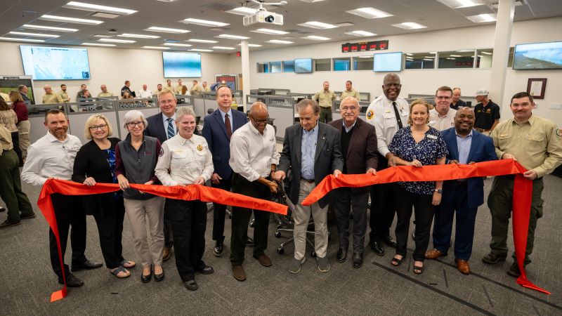Group of people holding a red ribbon. Middle two men are cutting ribbon with large scissors.