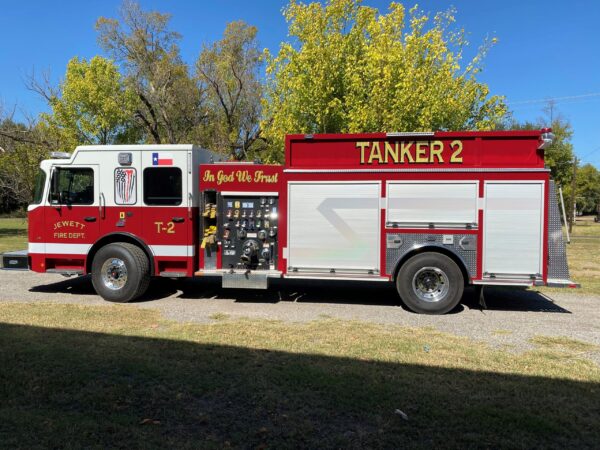 A red Jewett Fire Dept. tanker truck labeled "TANKER 2" with "In God We Trust" displayed, parked on grass with trees and a blue sky.