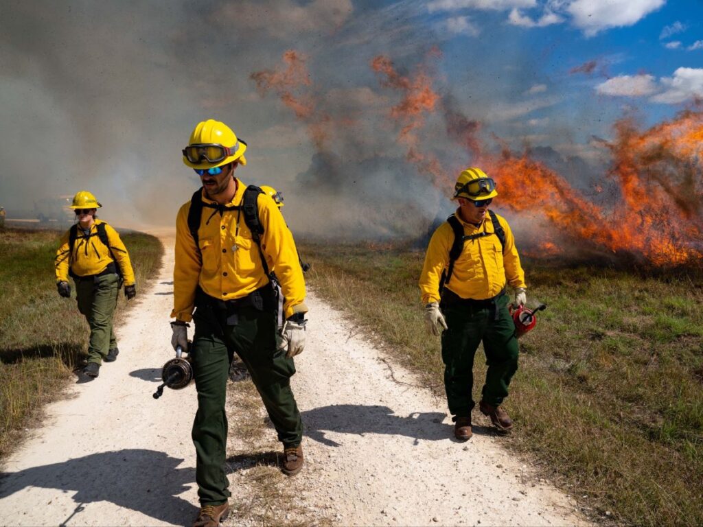 Tres bomberos sosteniendo torres de goteo alejándose de un incendio.