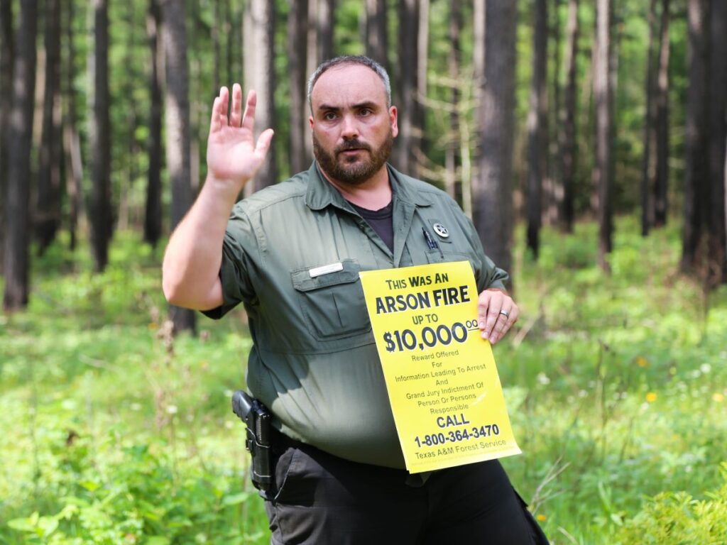 Law enforcement officer stands in forest and holds up yellow flyer for arson fire reward of up to $10,000