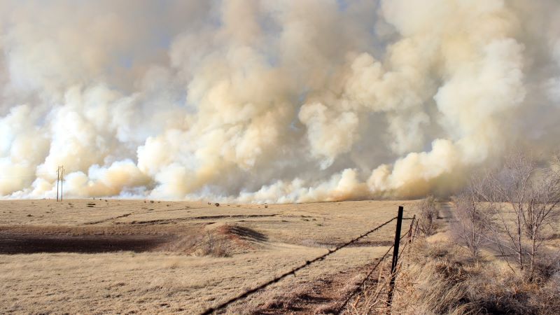 Fence along dry and tan field of grass with huge white and gray smoke plumes behind it