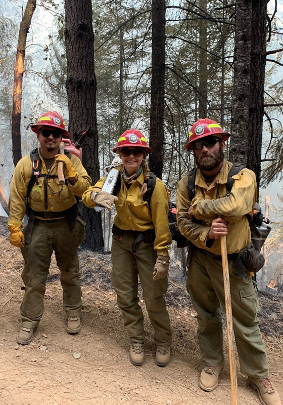Three firefighters with red hardhads stand in front of trees and smoke