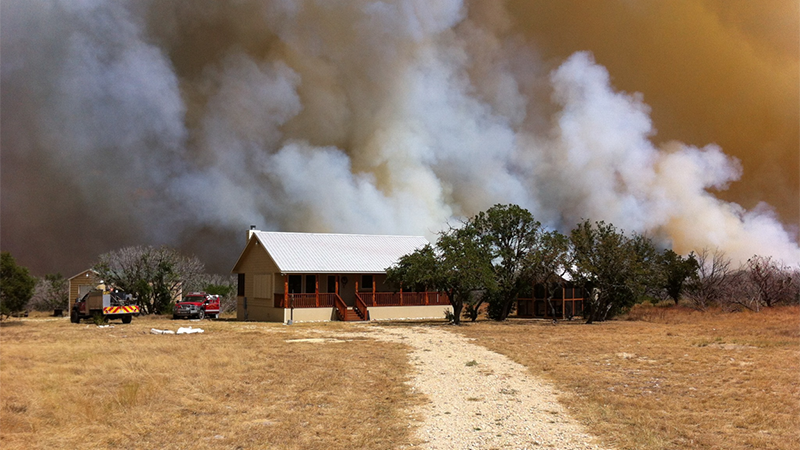 A house stands with a large wildfire and smoke behind it as firefighters respond.