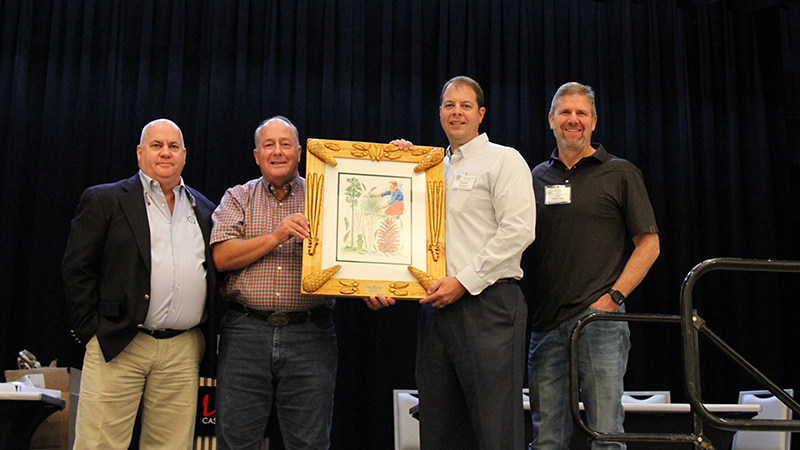 Four men with dress shirts stand on a stage in front of black background. Two men in the middle hold a wooden-framed painting of a pine tree and pine cone.