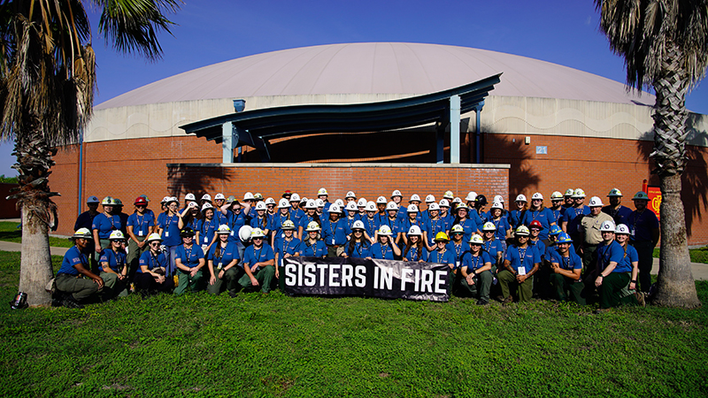 A large group of people wearing blue shirts and various hats, including hard hats and helmets, stand on a grassy area in front of a brick building with a dome-shaped roof. They hold a black banner with white text reading 'SISTERS IN FIRE,' framed by two palm trees. The image highlights unity and possibly women in firefighting or fire safety.