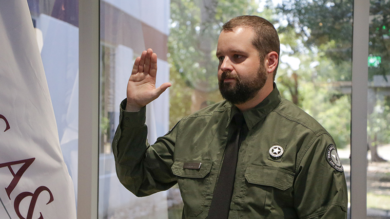 A Law Enforcement officer with a green shirt, black tie, and officer pin raises his right hand as he's sworn in.
