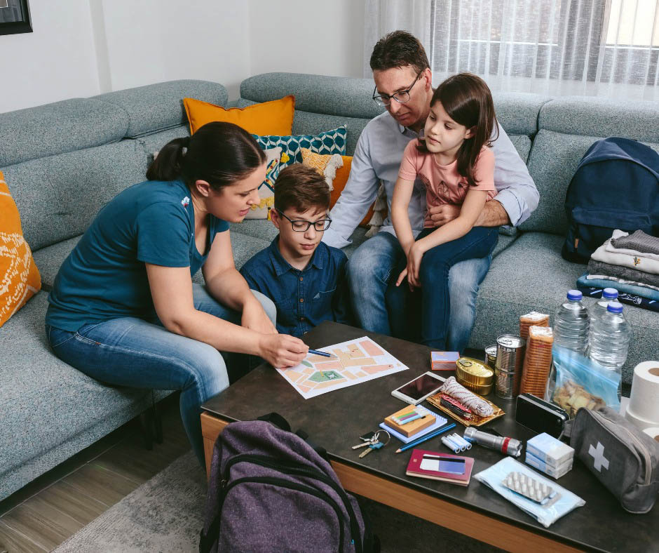 A family sits on a couch organizing emergency supplies including canned food, water bottles, and first aid items.